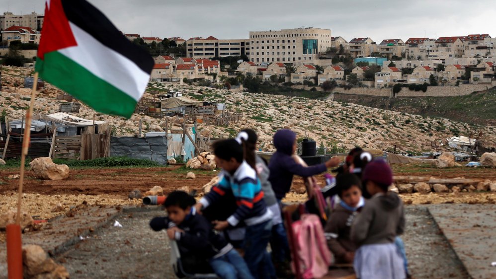 Palestinian kids playing in front of a Palestinian flag with an Israeli settlement in the background