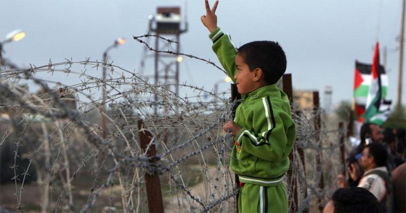 small palestinian boy standing next to a barbed wire fence with lookout tower in distance, holding his hand up in a peace sign