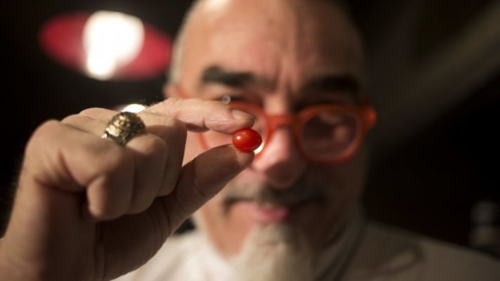 guy holding the small tomato between his fingers. the tomato is in focus in the foreground