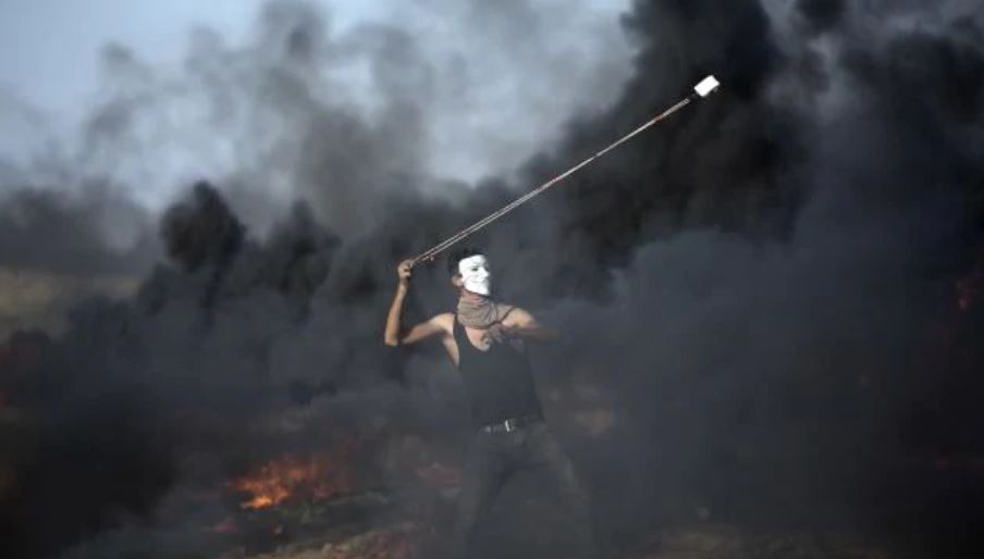 masked palestinian twirling a projectile around whilst smoke billows behind